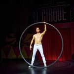 Bare-chested white brunet in white trousers stands in the middles of a large hoop, holding it up with one hand.