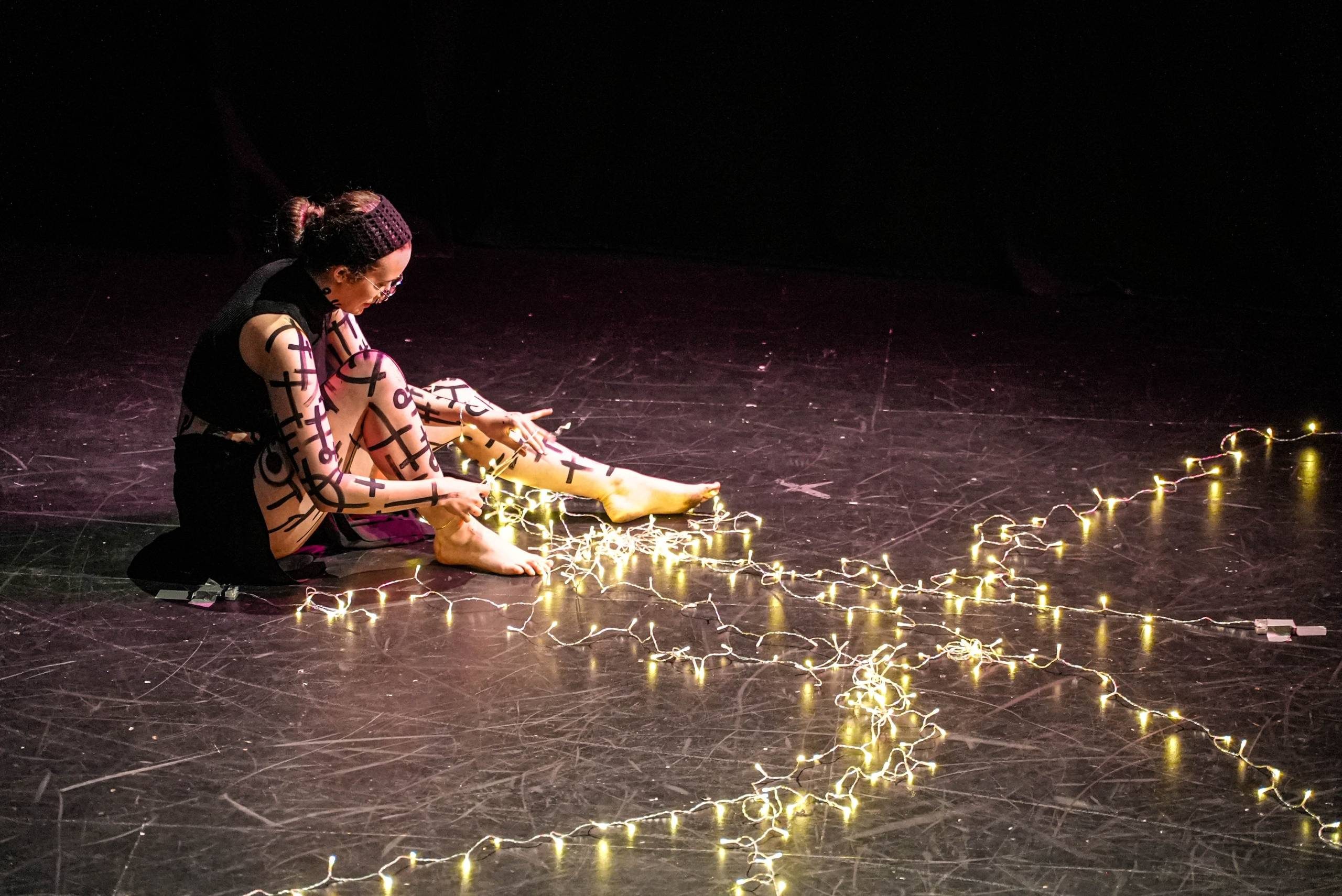 Artist Development A woman wearing all black with markings on her skin of lines and circles. She is surrounded by fairy lights on the stage.