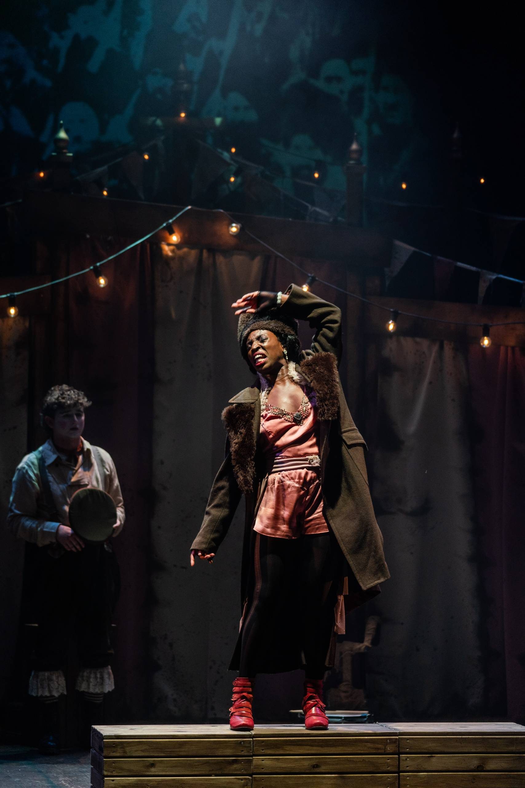 Oh What A Lovely War a female character stand on stage with disgust on her face while a man wearing clown makeup stands behind her watching.