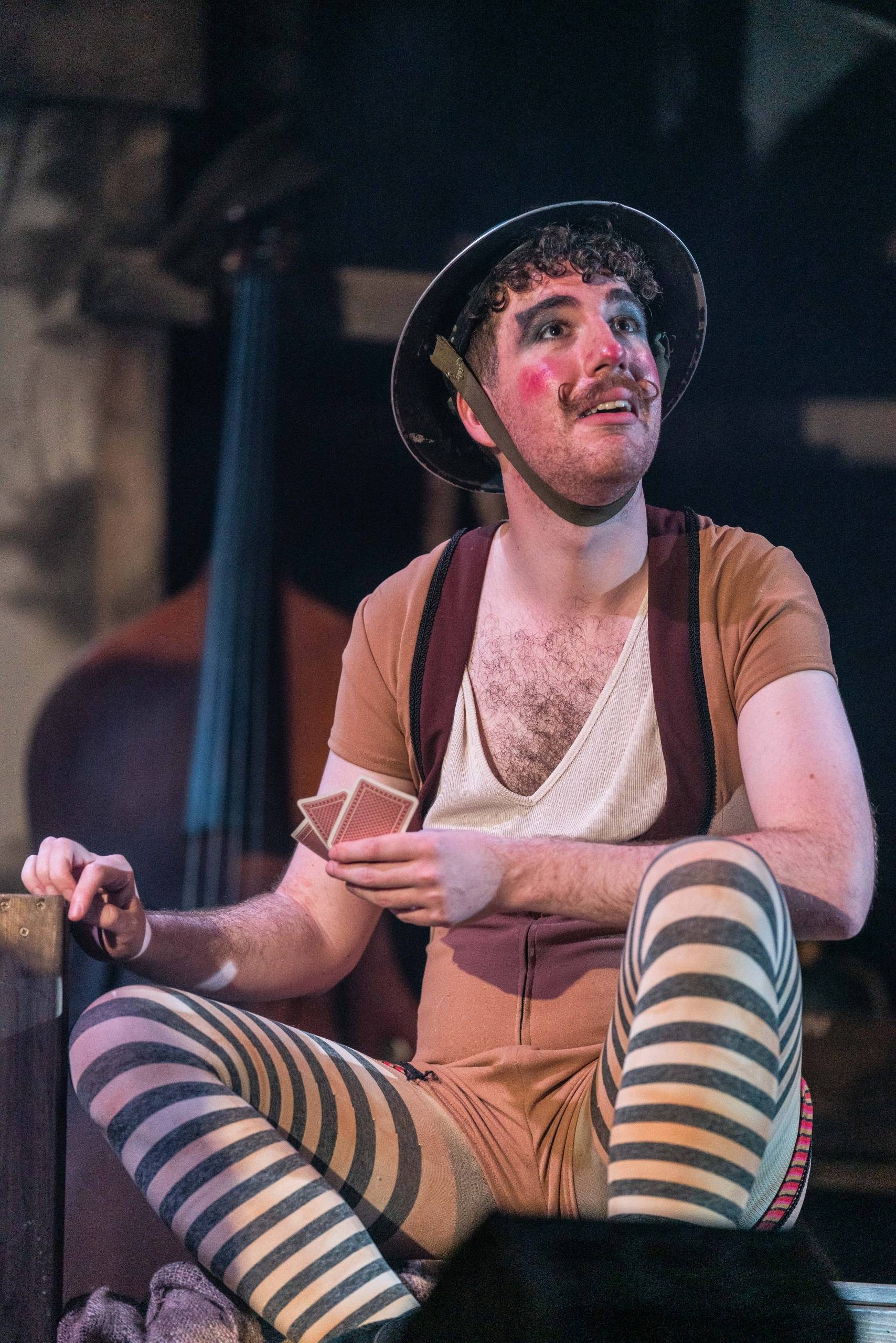 Oh What A Lovely War a man wearing stripy tights, brown top, face makeup and soldiers hat looks up.