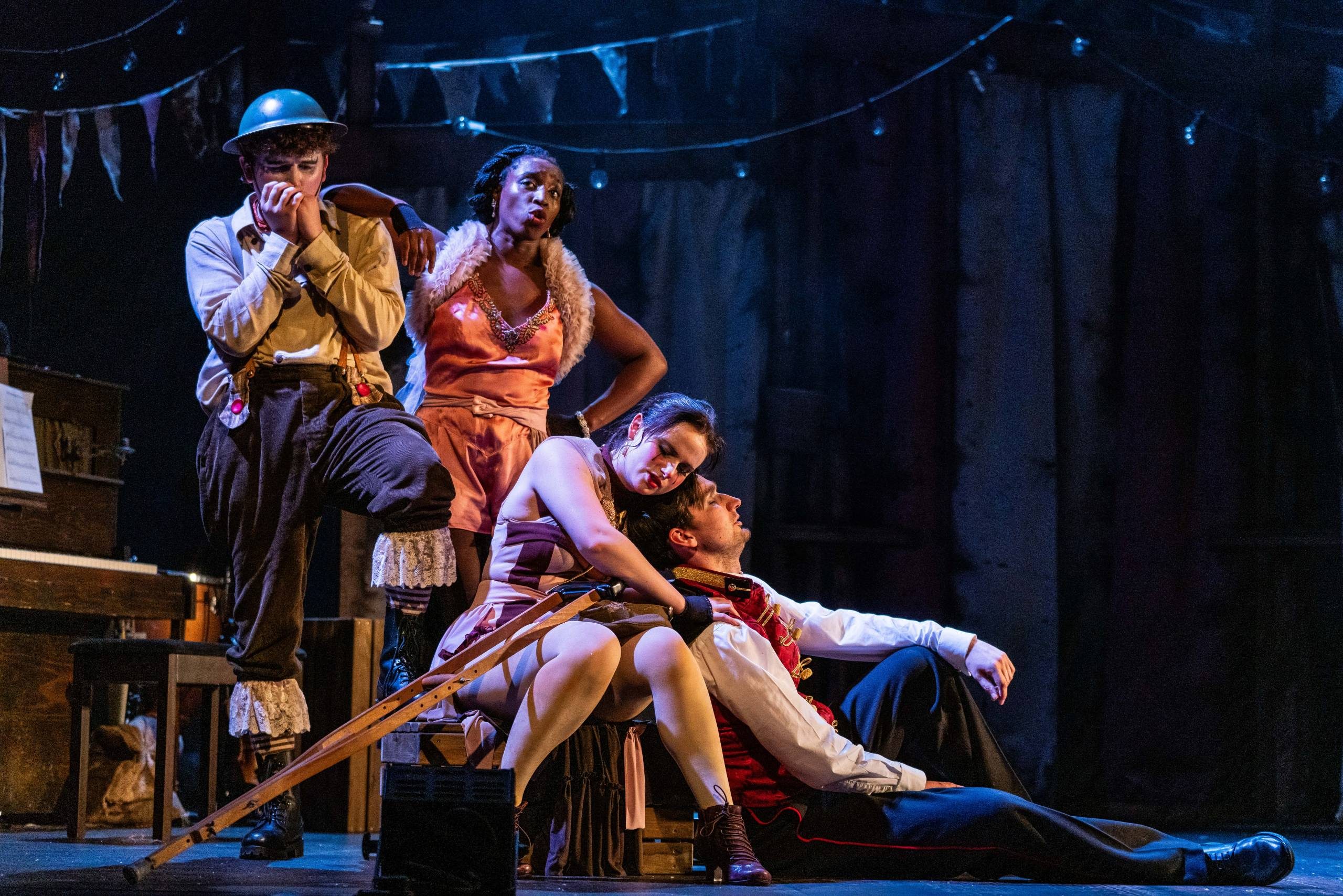 Oh What A Lovely War Four characters on stage, two male and two female. One male stands and plays the harmonica while one female has her arm on his shoulder. The other male and female sit on the floor together.