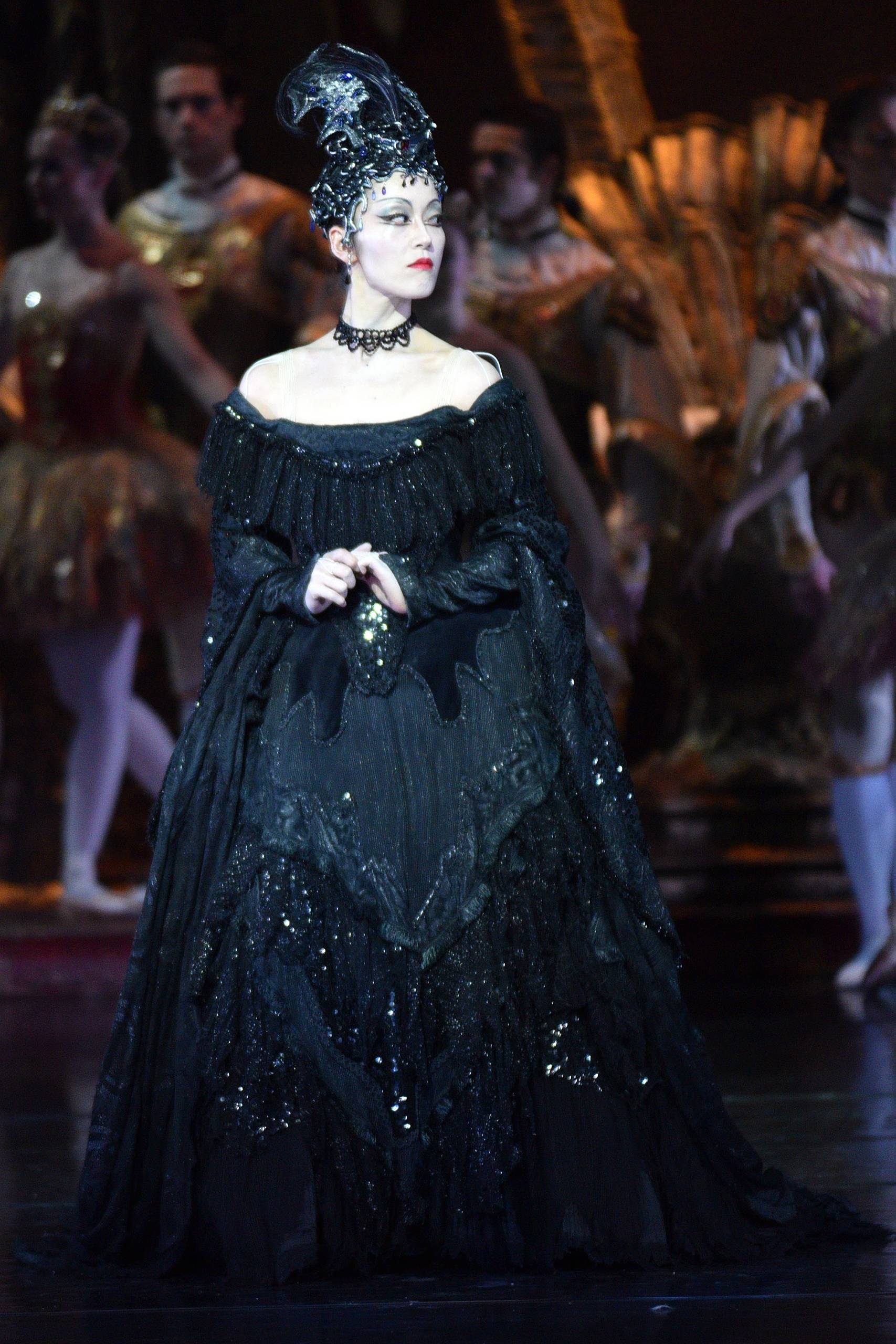 The Sleeping Beauty The evil queen stands tall and wears a long black dress. Ensemble dancers are in the background wearing white and gold costumes.