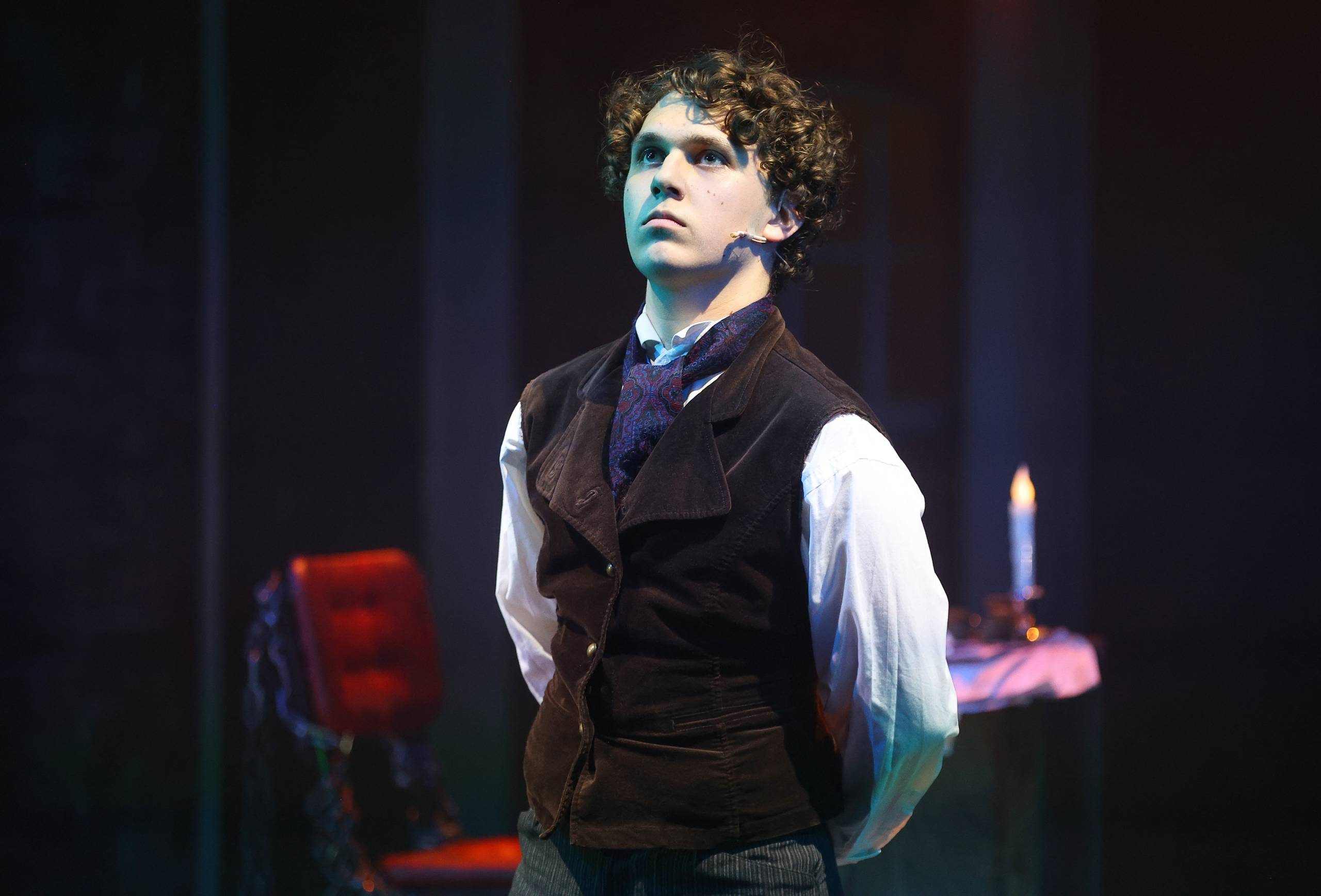 A Christmas Carol Scrooge stands proudly with his arms behind his back. Behind him is a red chair alongside a small table with a candle on top.