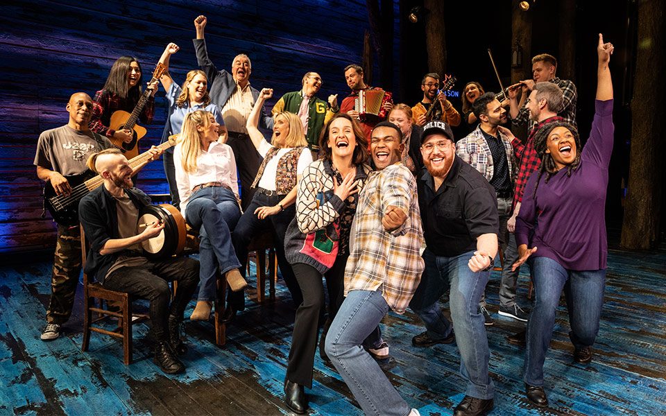 Come From Away A celebrating crowd punch the air. The back row play musical instruments