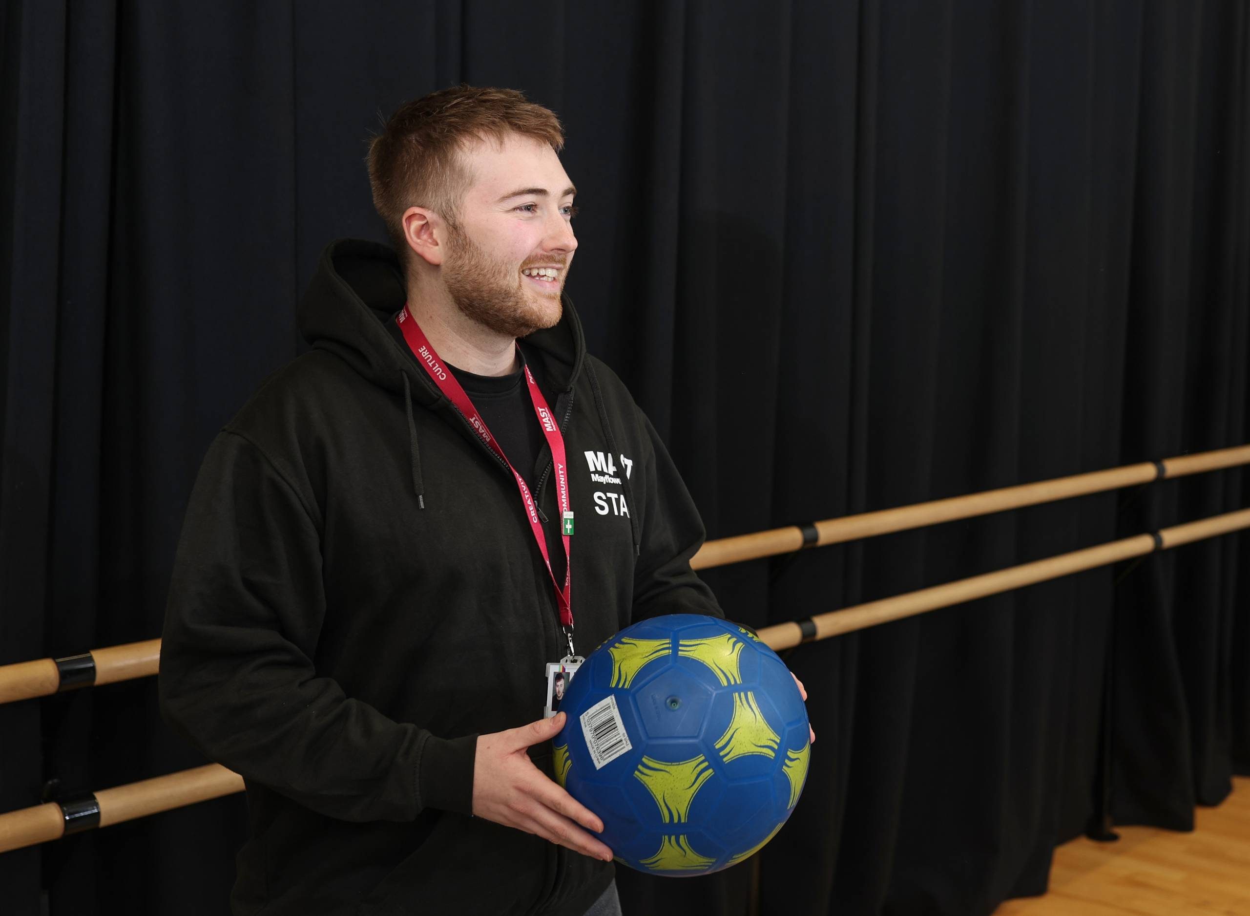 Workshops A man in a black MAST staff hoodie is holding a blue and yellow football and smiling to someone out of frame.