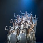 Matthew Bourne's Swan Lake Company Fourteen Swan-men flock on a bed, arms raised menacingly.