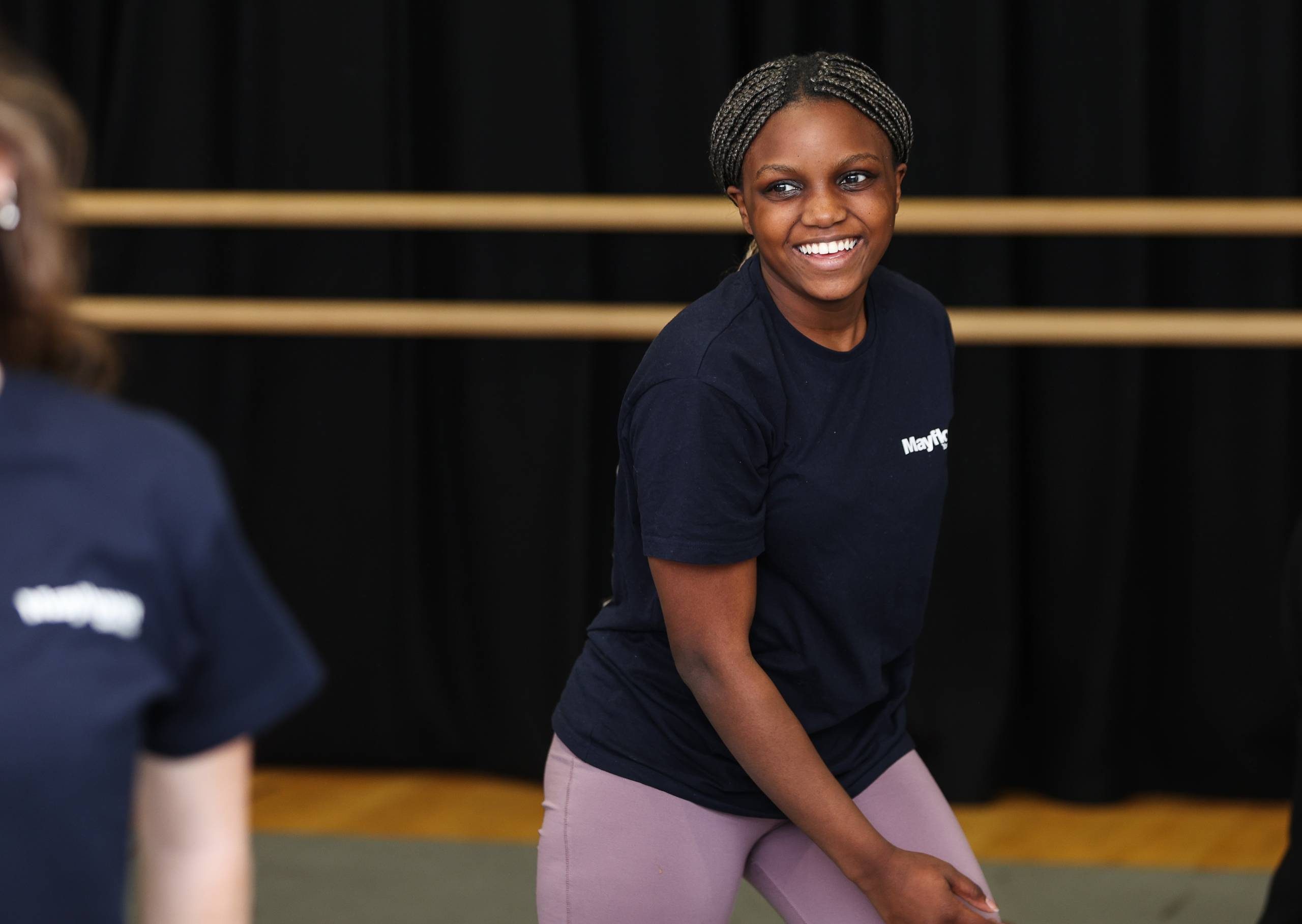 Mayflower Youth Theatre a young pariticpant wearing a navy blue Mayflower branded top and violet leggings is smiling.