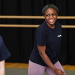 Mayflower Youth Theatre a young pariticpant wearing a navy blue Mayflower branded top and violet leggings is smiling.