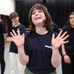 Mayflower Youth Theatre A young participant of our Youth Theatre is smiling and holding both hands in the air.