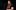 Residency image A girl with brown and pink hair speaking into a microphone on a stand. She is wearing a black t-shirt and is photographed from the waist up.