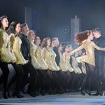 Line of dancers in gold and black holding hands as they watch couple twirling together face to face, woman in gold, man in black.