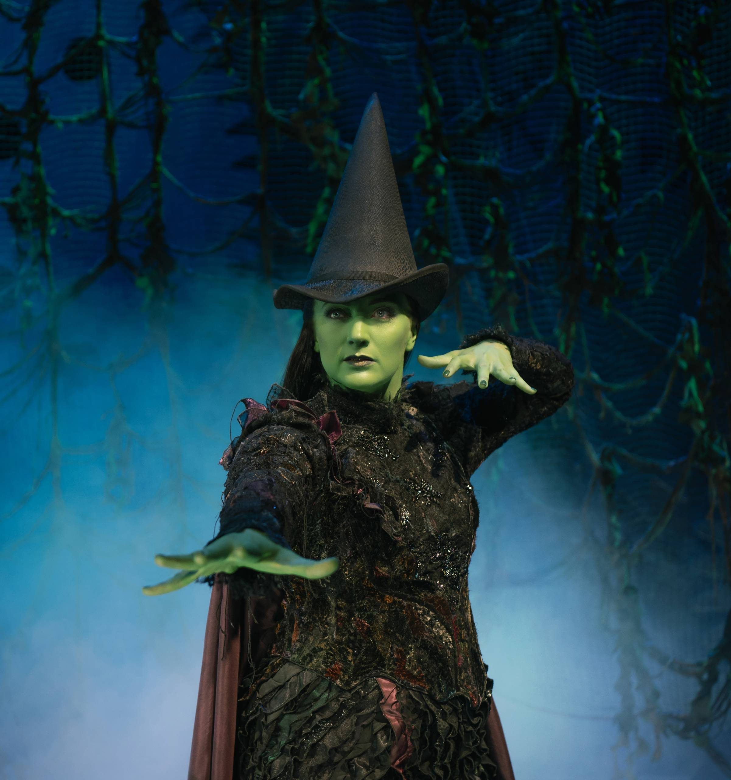Wicked Production image Laura Pick as Elphaba waving her hands as though casting a spell in a forest at night
