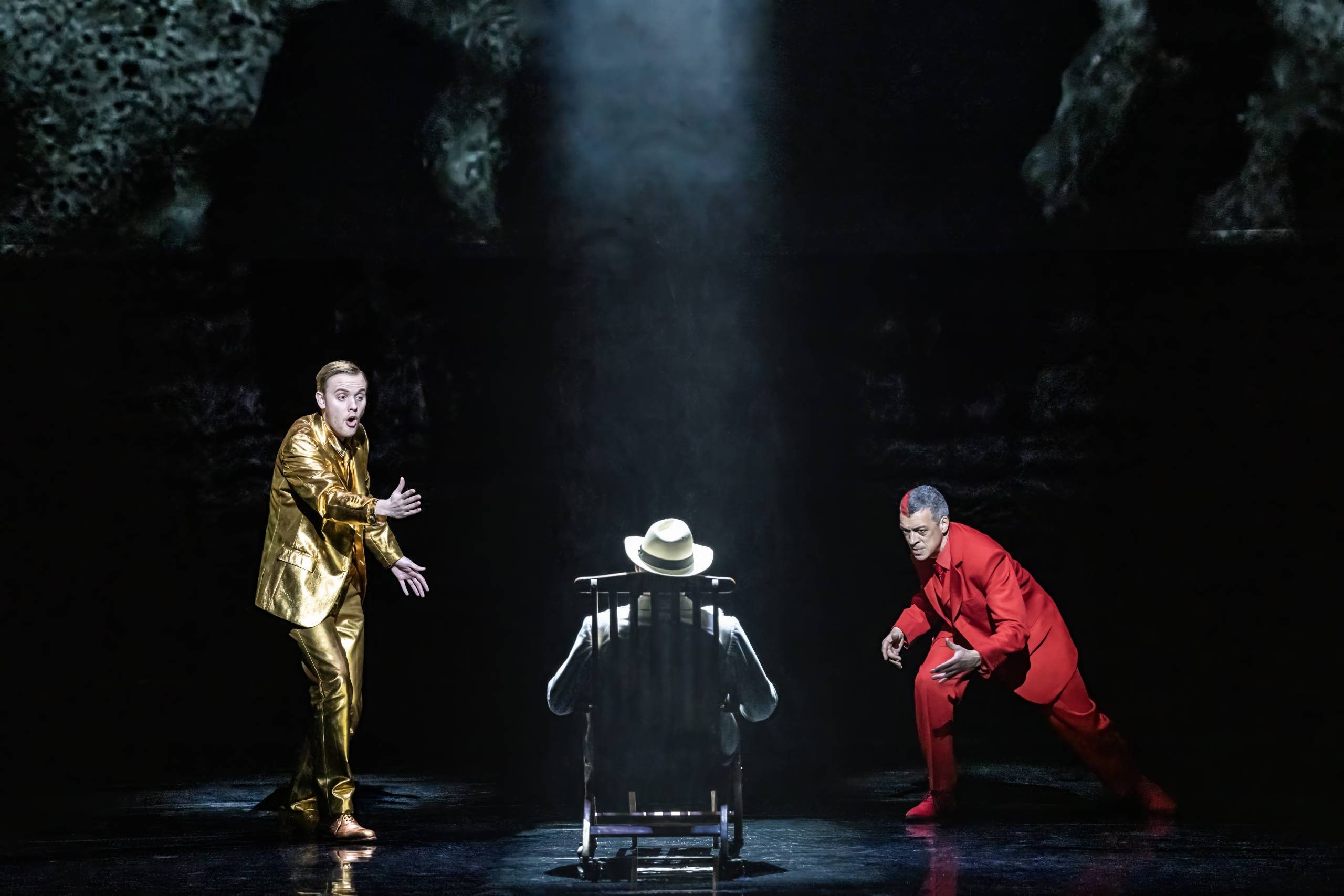 Death in Venice Dionysus and Apollo Apollo (Alexander Chance) in gold suit and Dionysus (Roderick Williams) in red suit, sing at Ashenbach facing away in wooden chair.