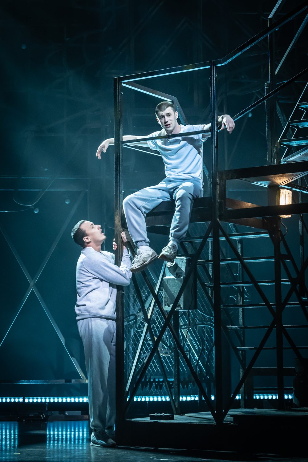 An Officer and a Gentleman production image Sid Worley (Paul French) looks up at Zack Mayo (Luke Baker) sitting on metal stairs, both wearing grey tracksuits